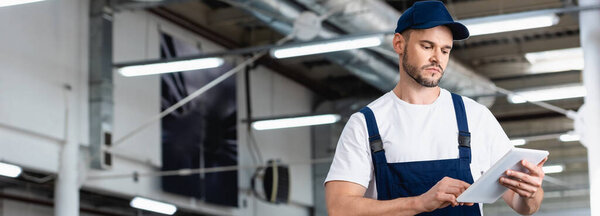 panoramic shot of mechanic in uniform and cap using digital tablet in workshop 