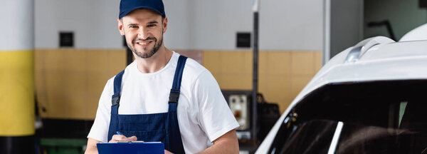 panoramic crop of mechanic in uniform holding clipboard near car