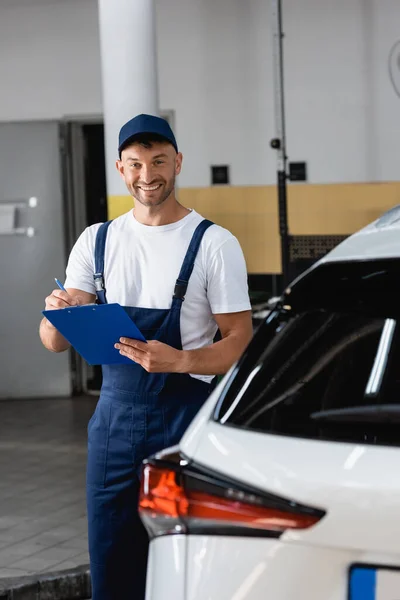 Vrolijke Monteur Cap Houden Klembord Buurt Van Moderne Auto — Stockfoto