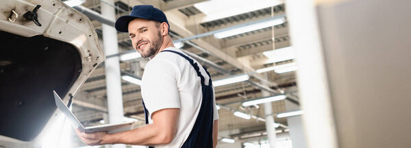 Panoramic shot of smiling auto mechanic holding laptop at service station