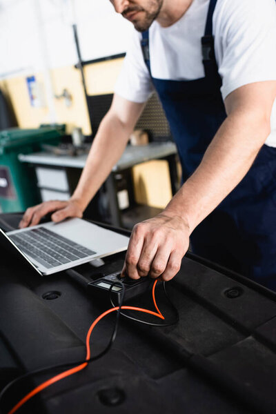 Auto mechanic leaning hands on car near laptop and turning lever of multimeter at service station