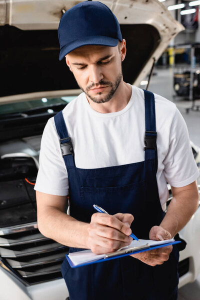 Auto mechanic writing on clipboard near open hood of car at service station