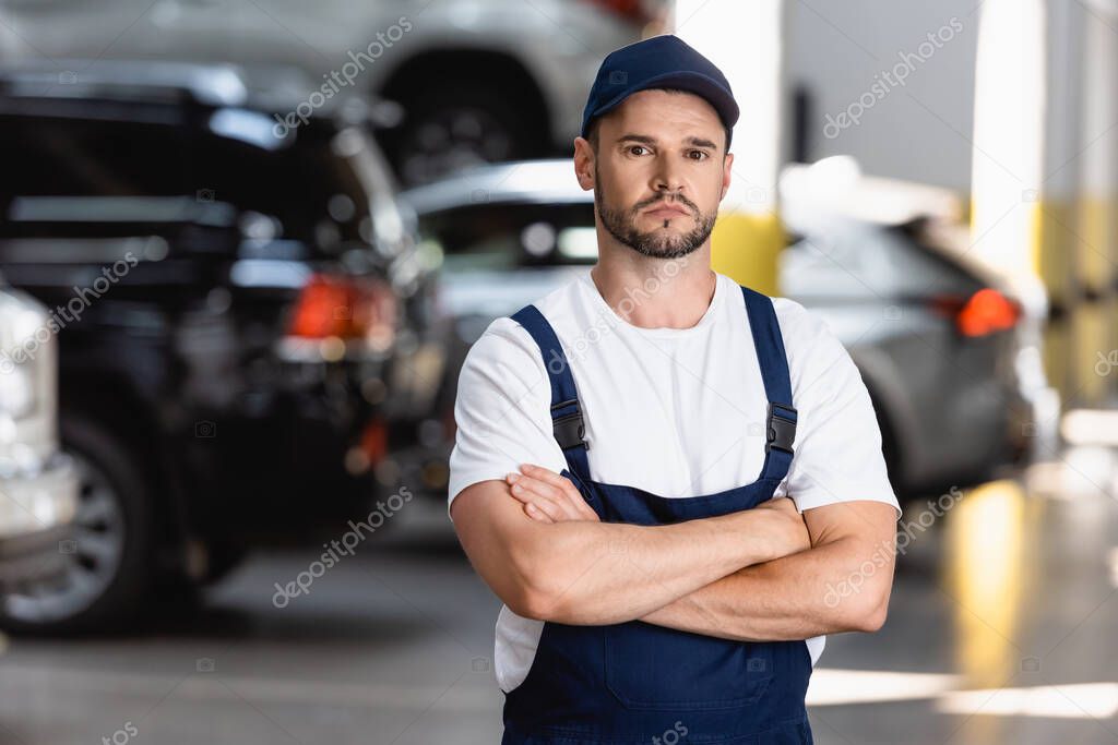 handsome mechanic in uniform and cap standing with crossed arms in car service