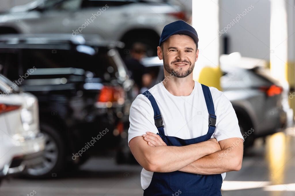 happy mechanic in uniform and cap standing with crossed arms in car service
