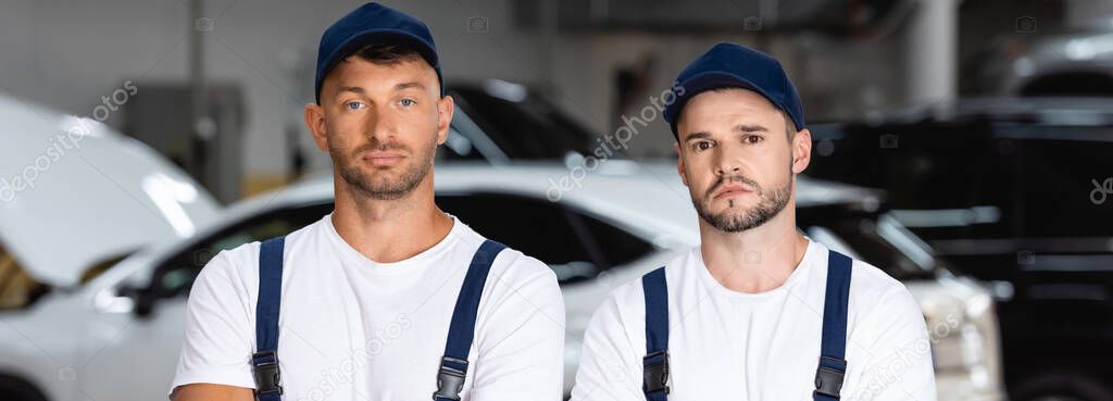 panoramic concept of handsome mechanics in caps looking at camera in car service