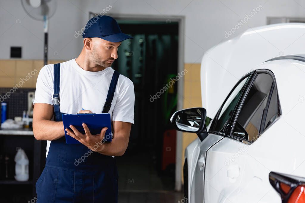 handsome mechanic in cap holding clipboard and looking at car