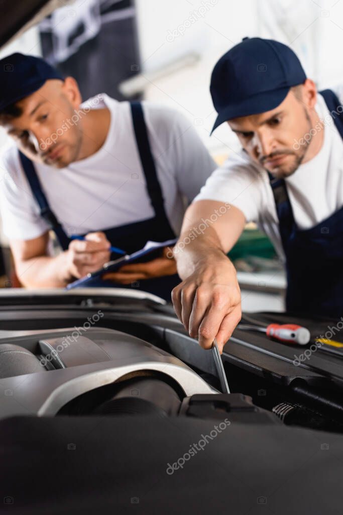 selective focus of mechanic in uniform repairing car near coworker with clipboard and pen 