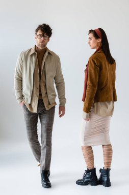 full length view of trendy couple posing on grey while man looking at camera on grey clipart