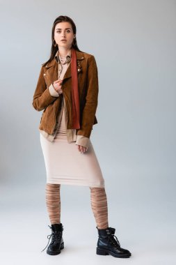 full length view of attractive girl in trendy autumn outfit touching suede jacket on grey clipart