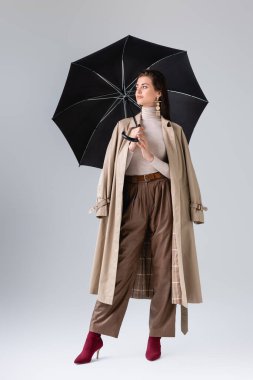 Gri üzerinde siyah şemsiyeyle poz veren zarif bir kadının tam boy görünümü