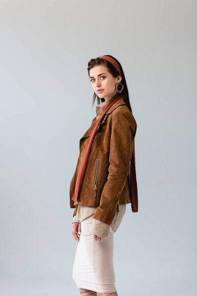 attractive, fashionable girl in suede jacket looking at camera isolated on grey