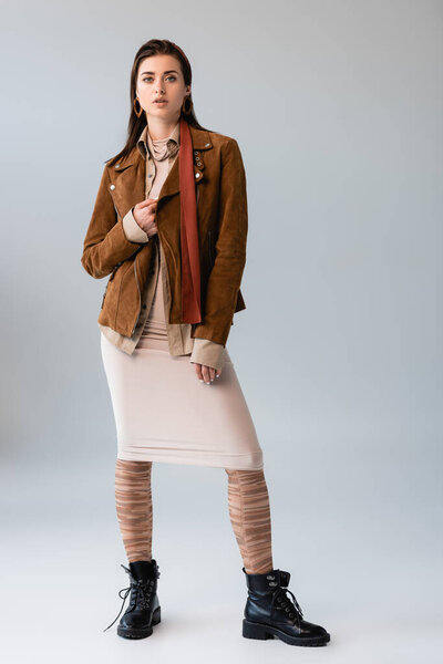 full length view of attractive girl in trendy autumn outfit touching suede jacket on grey