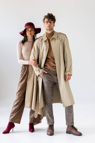 full length view of stylish woman in hat touching hand of trendy man in trench coat on grey