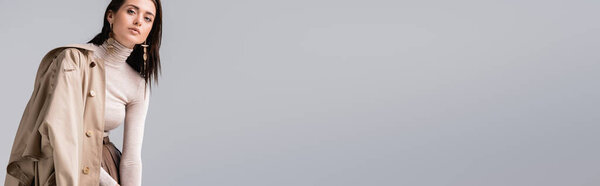 горизонтальное изображение стильной девушки в плаще на плече, смотрящей на камеру, изолированную на сером