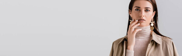 горизонтальное изображение красивой, элегантной девушки, касающейся лица при взгляде на камеру, изолированную на сером