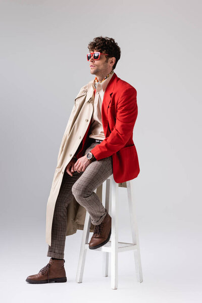 handsome, stylish man with trench coat on shoulder sitting on stool and looking away on grey