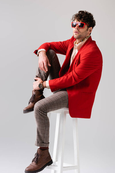 fashionable man in red blazer and sunglasses touching leg while sitting on stool isolated on grey