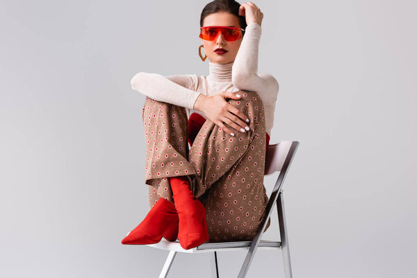 Привлекательная девушка со скрещенными ногами, держась за руку возле головы и сидя на стуле, изолированном на сером 