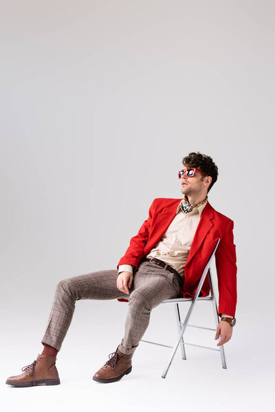 Stylish man in red blazer leaning back and posing while sitting on chair on grey