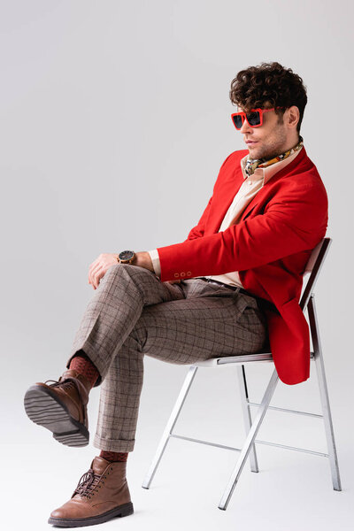 Stylish man in red blazer with crossed legs, sitting on chair on grey