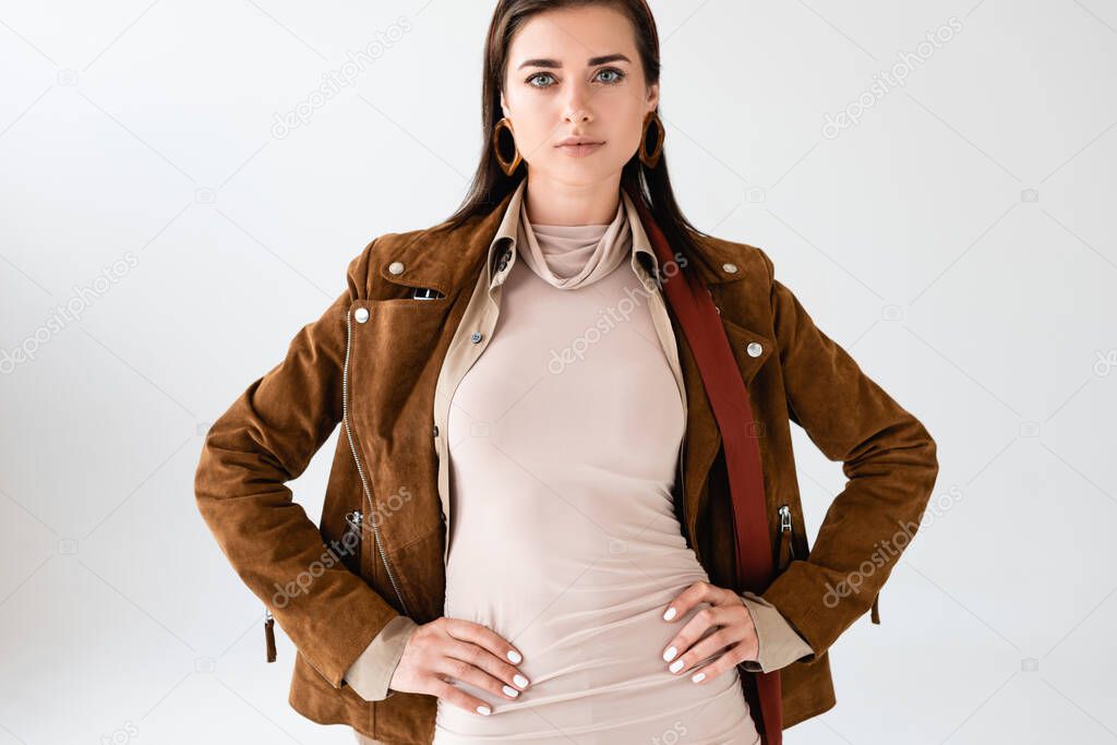 confident, trendy girl in suede jacket posing with hands on hips isolated on grey