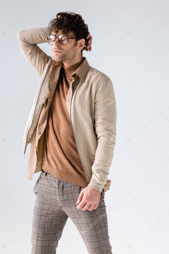 trendy man in eyeglasses touching hair while posing with closed eyes isolated on grey