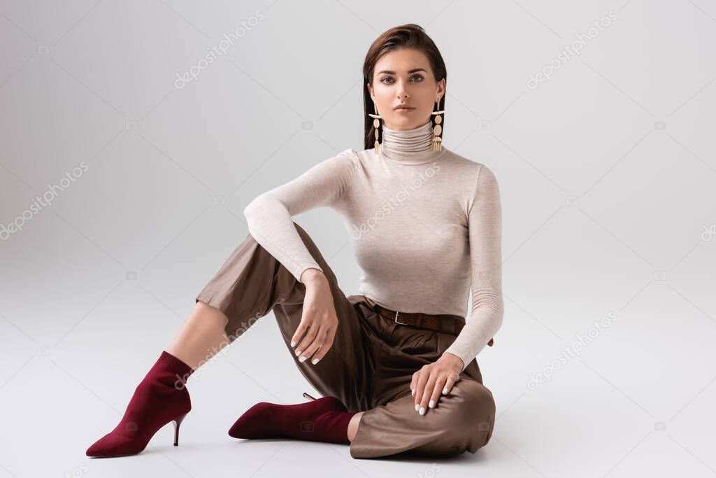 attractive, stylish woman sitting on floor and looking at camera on grey