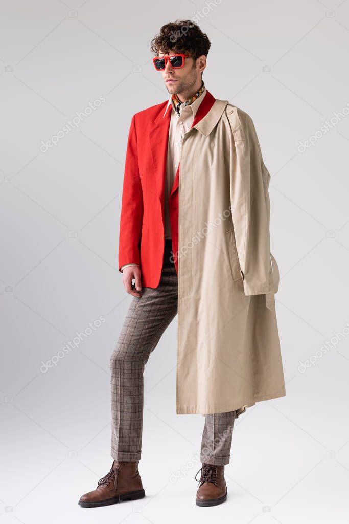 full length view of trendy, confident man with trench coat over shoulder looking away on grey