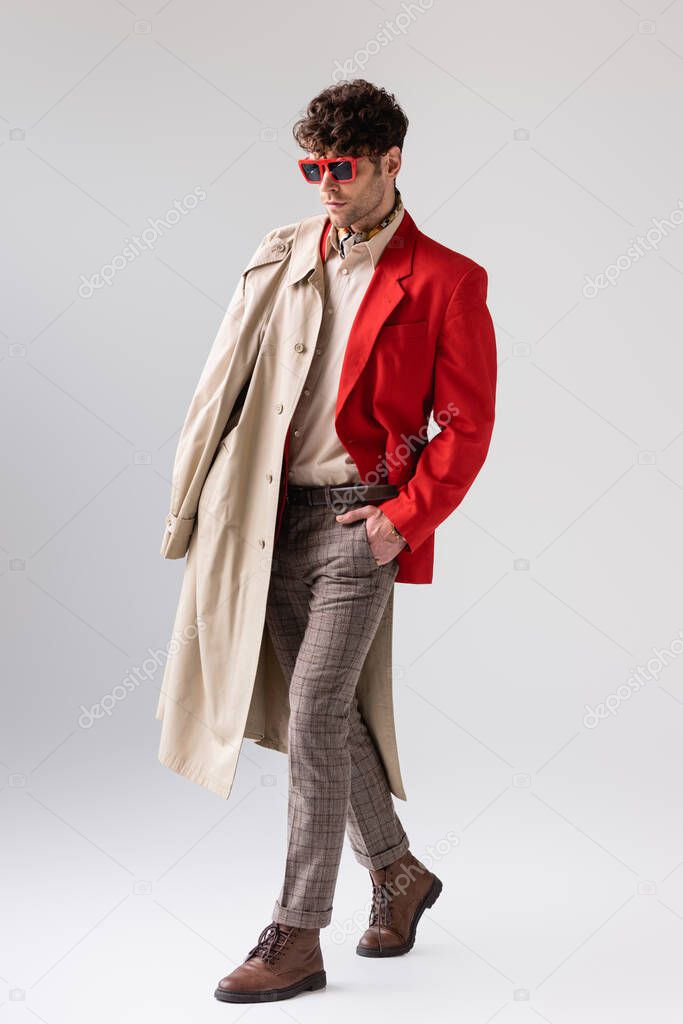 full length view of trendy man with trench coat over shoulder posing on grey with hand in pocket