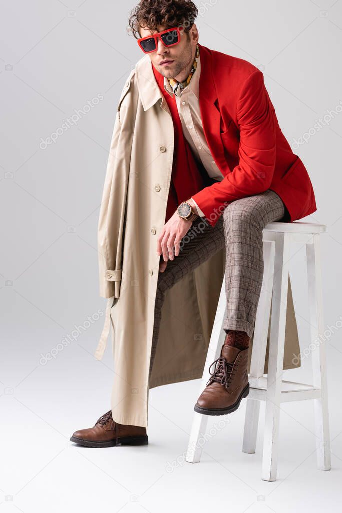 fashionable man in red blazer with trench coat on shoulder sitting on grey