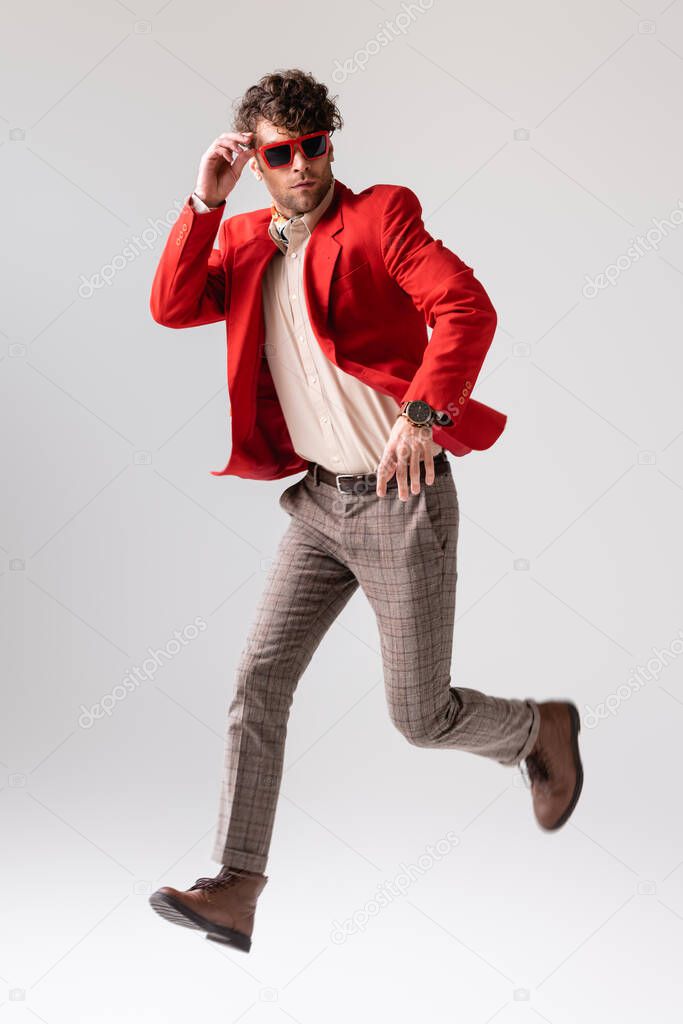 full length view of trendy man touching sunglasses and looking back while running on grey