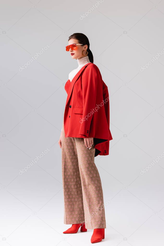 full length view of beautiful, stylish girl with red blazer on shoulder looking away on grey