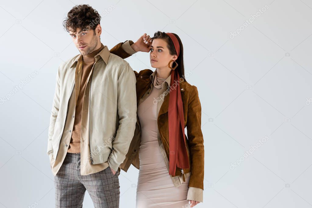 Trendy girl in autumn outfit posing near stylish man with hands in pocket isolated on grey 