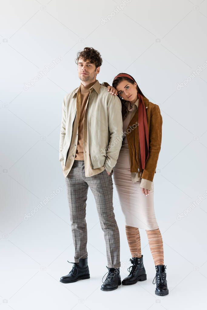 Full length view of stylish woman in autumn outfit leaning on man standing with hands in pockets on grey
