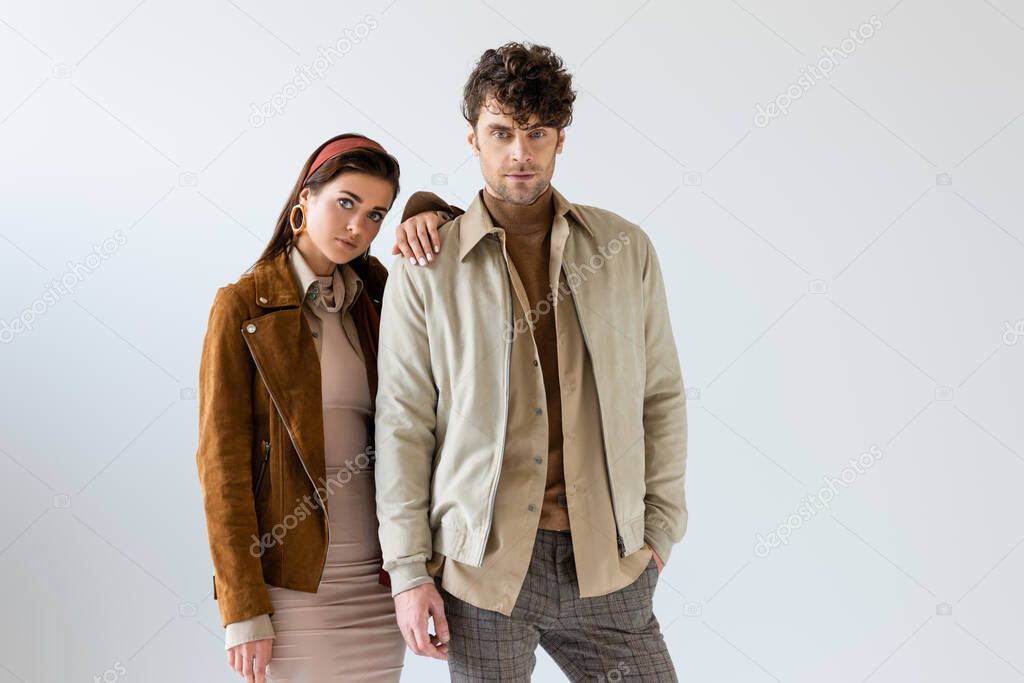 Attractive girl in autumn outfit leaning on handsome man standing with hand in pocket on grey