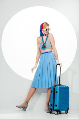 Güneş gözlüklü, gülümseyen stil sahibi kadın beyaz arka planda mavi bavul tutuyor.