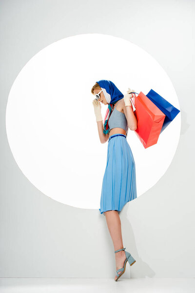 Вид сбоку на триумфальную женщину, держащую красные и синие сумки для покупок возле круглой дыры на белом фоне 