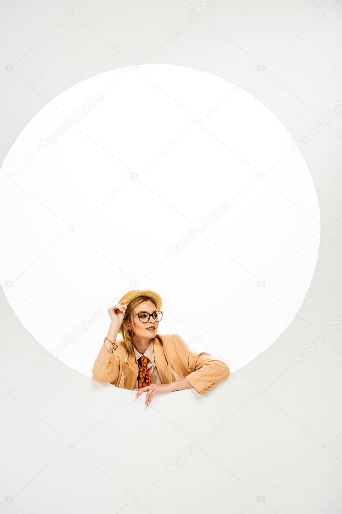 Trendy woman in straw hat and eyeglasses looking away beside round hole on white background 