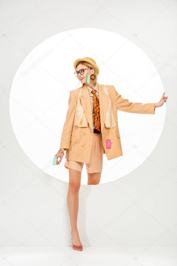 Fashionable woman with price tags on clothes smiling beside circle on white background 