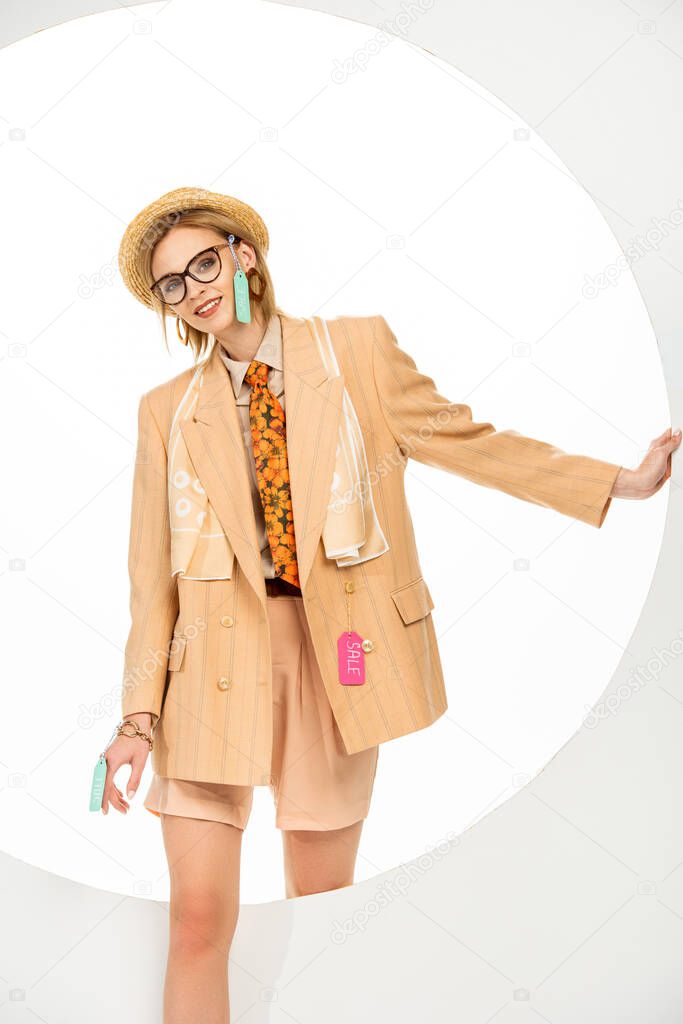 Stylish girl in clothes with price tags smiling at camera near round hole on white background 