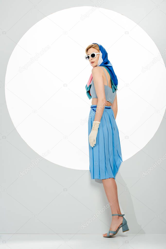 Side view of trendy woman in blue skirt and sunglasses standing near circle on white background 