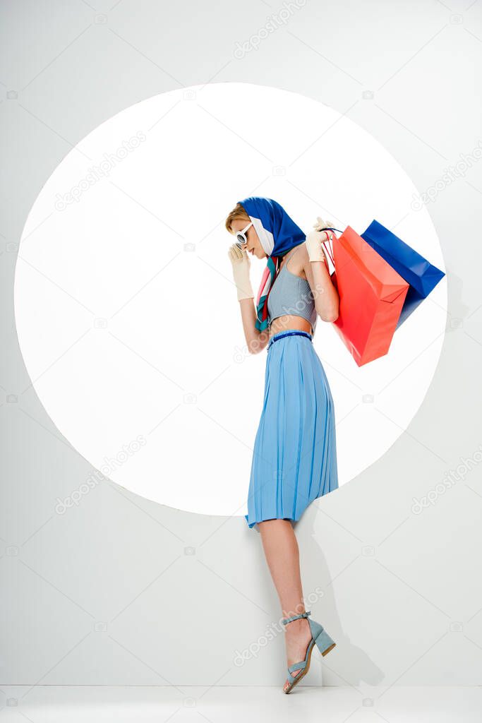 Side view of trendy woman holding red and blue shopping bags near round hole on white background 
