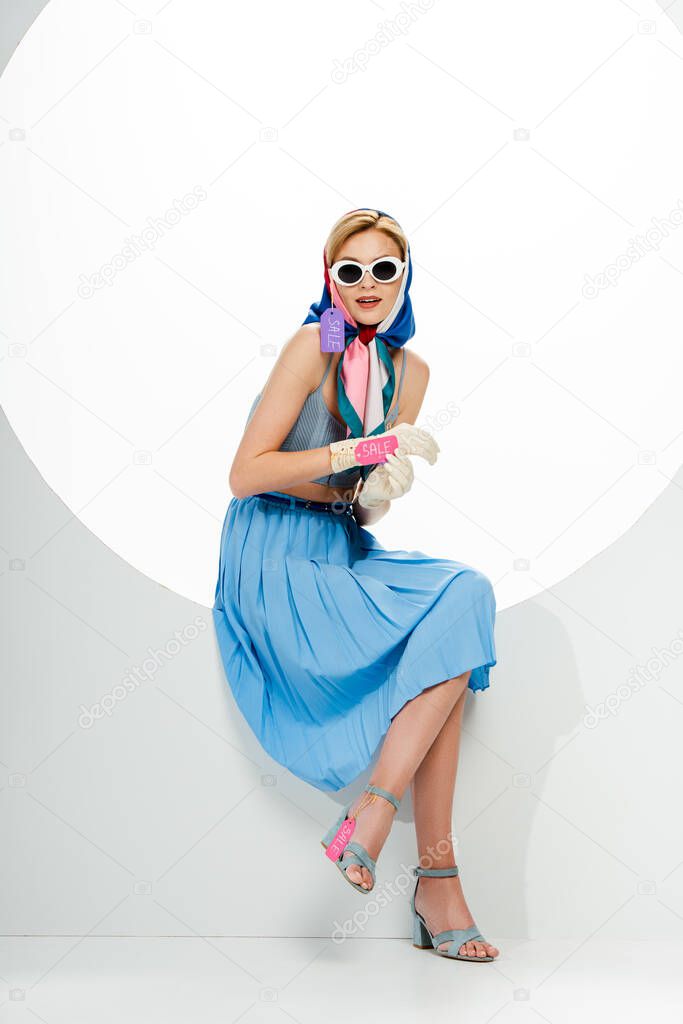 Fashionable young woman with price tags with sale lettering sitting in round hole on white background 