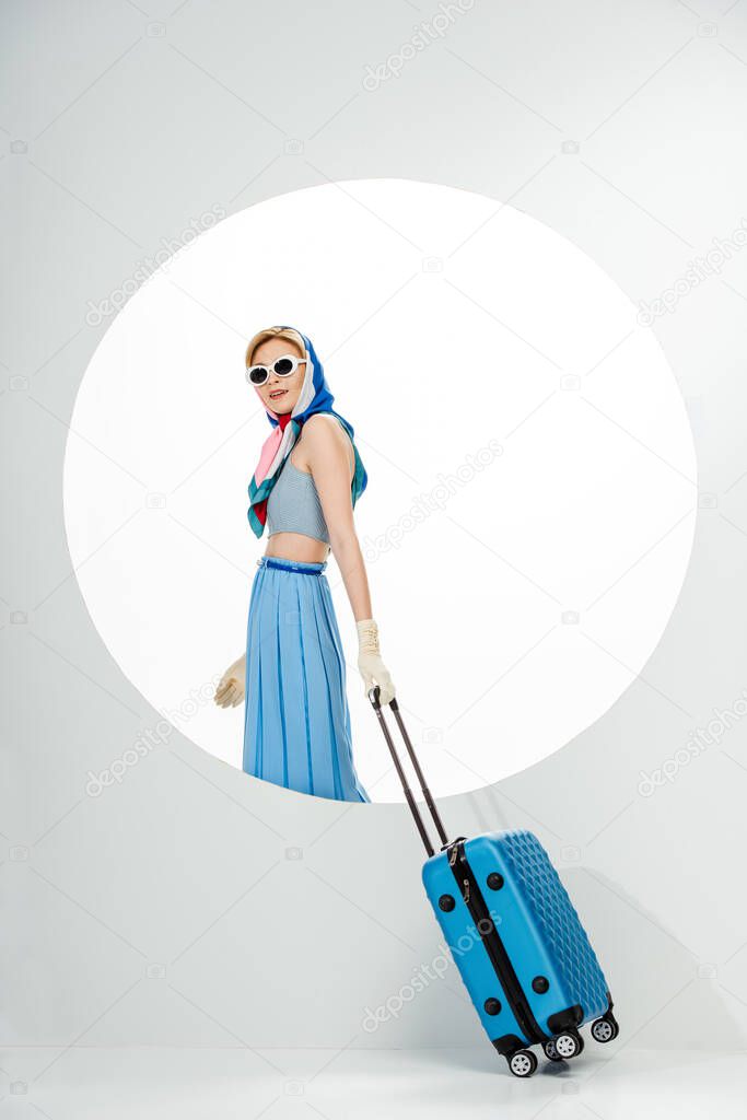 Stylish woman in sunglasses holding blue suitcase near circle on white background 