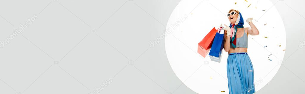 Horizontal image of positive stylish girl holding red and blue shopping bags under falling confetti near circle on white background