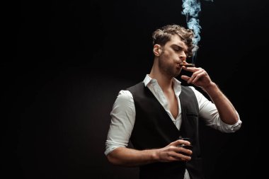 Gömlek ve yelek giyen, puro içen ve siyah üzerine izole edilmiş viski bardağı tutan adam. 