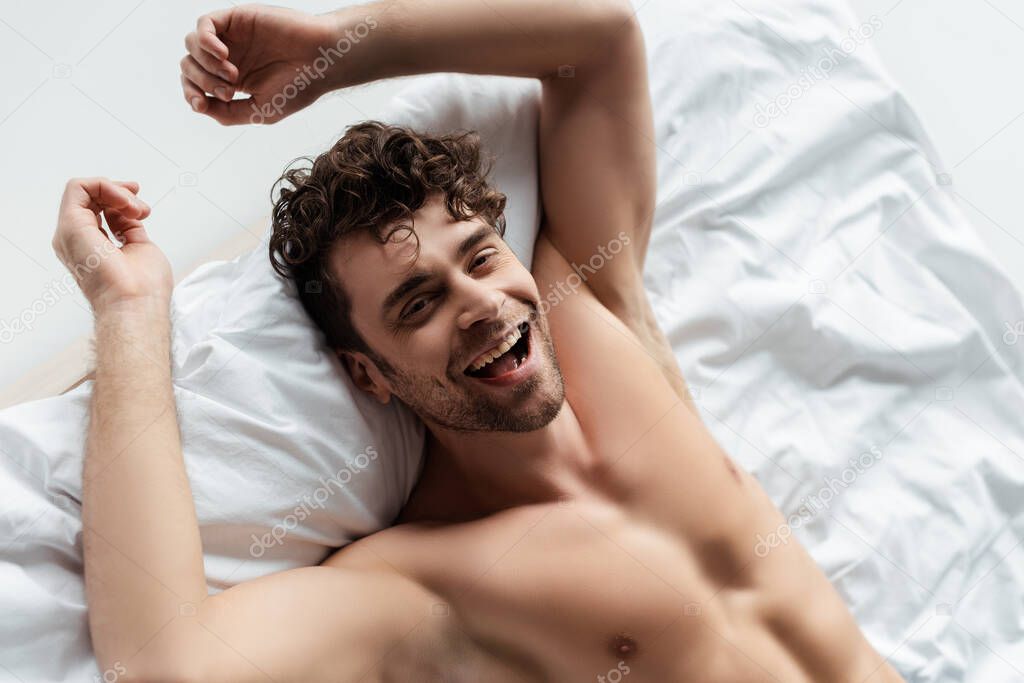High angle view of cheerful shirtless man smiling at camera on bed isolated on white