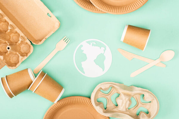 Top view of paper earth sign near disposable tableware and wooden cutlery on green background, ecology concept