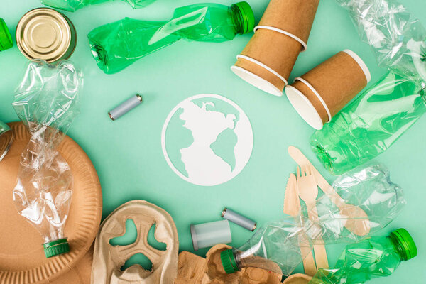 Вид сверху на пластиковые бутылки, аккумуляторы и одноразовую посуду возле знака "бумажная земля" на зеленом фоне, концепция экологии