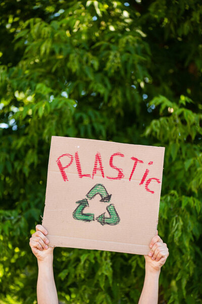 Cropped view of man holding placard with plastic lettering and recycle sign with trees at background, ecology concept 